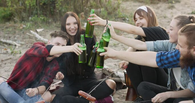 Close view of friends frying sausages sitting around bonfire, drinking beer on sandy beach. Young group of men and women with beverage singalong playing guitar near campfire