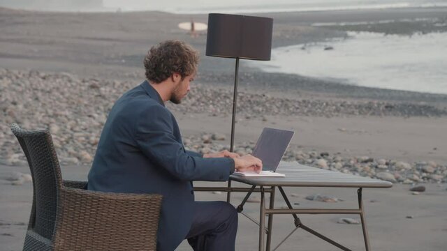 Wide side-view medium shot of businessman in blue jacket sitting at desk in front of blue ocean working on laptop