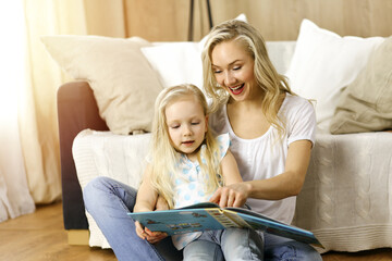 Happy family. Blonde young mother reading a book to her cute daughter while sitting at wooden floor in sunny room. Motherhood concept