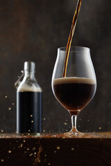 Stream of dark stout pours from the bottle into a beer glass with splashes on a wooden table. Jet of the dark porter pours and whips the thick foam in a half-pint glass. Selective focus