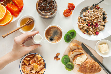 Winter breakfast with dried fruit granola and candied fruit on a white table. A woman's hand holds a milkman over a cup of fresh coffee. View from above