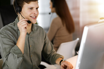 Young dark-haired guy with headsets is talking to a client, while sitting in sunny office. Call center operators at work