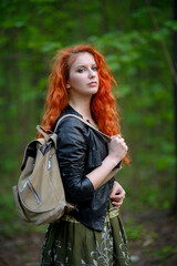 Fototapeta na wymiar Pretty young sexy fashion sensual red hair woman posing in a forest background dressed in hipster rock style outfit, leather jacket and tourists backpack