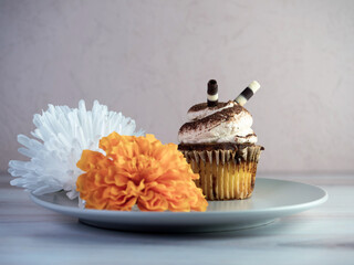 Tiramisu cupcake with white frosting and espresso powder with chocolate sticks sitting on a gray plate with an orange and white flower bloom.  Delicious coffee flavored dessert.