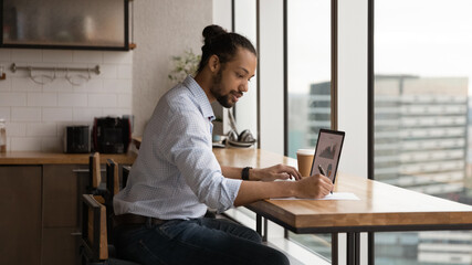 Focused young biracial man sales manager prepare summary report at home office check stats on pc screen make records on paper. Black male financial expert study stock prices rate growth graphs online