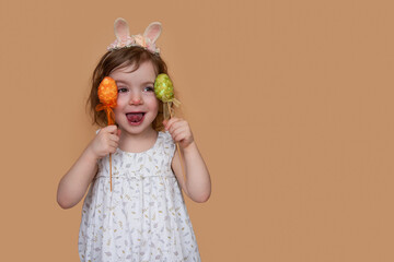 Positive portrait of little girl with rabbit ears on her head, showing her tongue holding an orange green egg. Isolated light orange background, copy space, postcards. Festive child in costume bunny