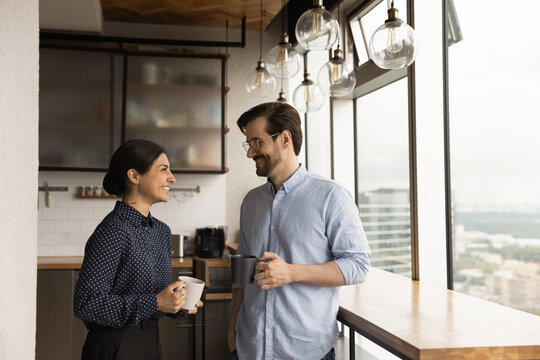 Happy mixed race family couple drink tea coffee at new modern apartment flat by picture window. Loving young spouses indian wife caucasian husband enjoy talking holding cups with hot drinks at kitchen