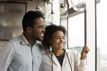 Happy afro american husband wife new studio apartment owners stand by panoramic window hold keys...