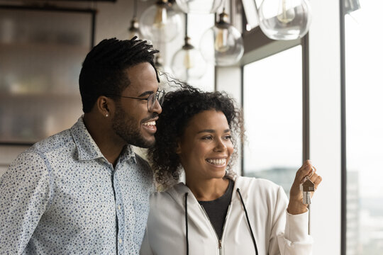 Happy afro american husband wife new studio apartment owners stand by panoramic window hold keys dreaming. Excited young black spouses becoming homeowners enjoy splendid city picture from modern flat