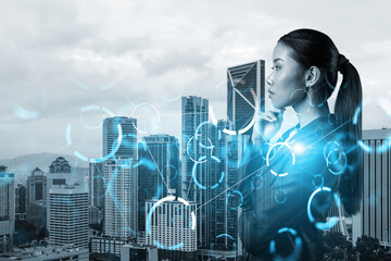 Attractive young Asian businesswoman pondering on technology at business process to achieve tremendous growth in commerce. Tech hologram icons over Kuala Lumpur background - Powered by Adobe