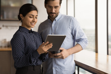 Young hindu female office employee mentor show interested male intern problem solution online using tablet. Two coworkers of different ethnicities discuss work meeting at dining area on lunch break.