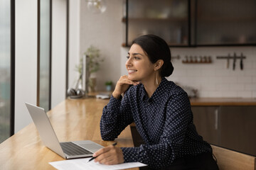 Inspired mixed race female sit by table with laptop at home kitchen look at window engaged by...