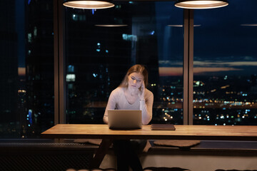 Unhappy young lady need to stay at modern office with window wall after workday to finish task. Sad...