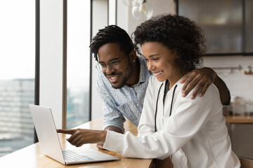 Happy smiling black couple in love cuddle at kitchen choose goods services online on laptop screen....