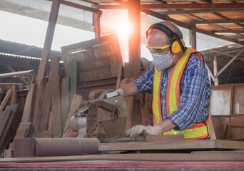 Elderly carpenter craftsman wearing safety gear working with hardwood using a miter saw to cut a piece of wood in the workshop. Manufacture of wood products..