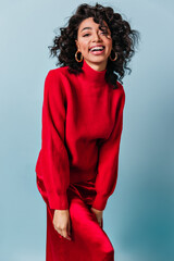 Blissful curly woman posing on blue background. Front view of blithesome girl in red sweater.