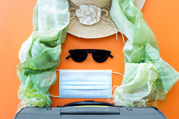 Flat lay with copy space, a luggage, face mask, sun hat, sunglasses and a scarf are arranged on an orange background. Travel concept during the Covid-19 pandemic.