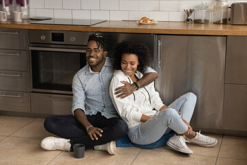 Affectionate happy african spouses cuddle on floor at modern kitchen interior relaxing enjoying good time together. Stylish young black couple in love having date at home celebrate moving to new house