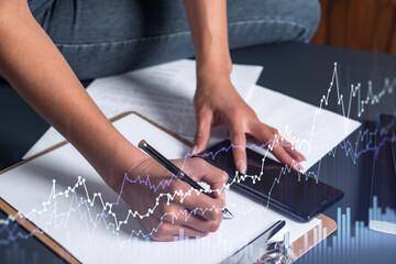 A woman trader in casual wear writing down some quotes to research stock market trends using smart phone for right investment. Wealth management concept. Hologram Forex chart over close up shot.