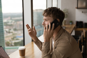 Stylish young man in casual clothing engaged in business phone talk look at beautiful urban panorama through large window. Millennial businessman hold cell at ear discuss new project with colleague