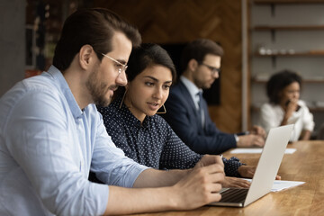 Focused millennial male intern look at laptop screen listen to skilled indian female mentor. Hindu woman experienced worker consult young man new employee help in computer work at corporate network