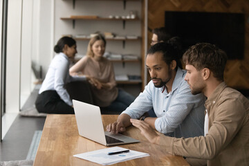 Two diverse male employees colleagues sit by laptop in modern office area engaged in working on project startup together. Capable young man share idea experience with black guy intern trainee using pc