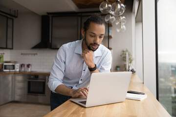 Thoughtful young african male stand by desk with laptop at home kitchen read important message email think on answer create respond. Focused biracial man student contact friend using messenger pc app - Powered by Adobe