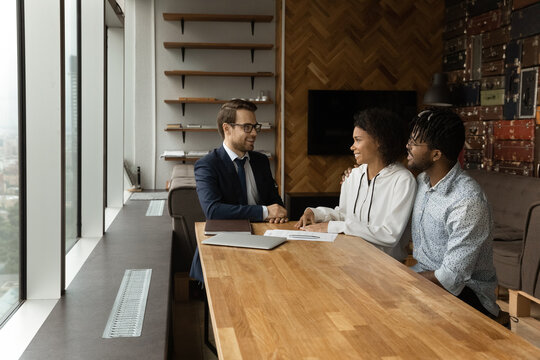 Happy African American Couple Sit By Desk At Travel Agent Office Listen To Pleasant Male Consultant Choose Trip. Confident Young Businessman Talk With Black Husband Wife Customers Offer House To Buy