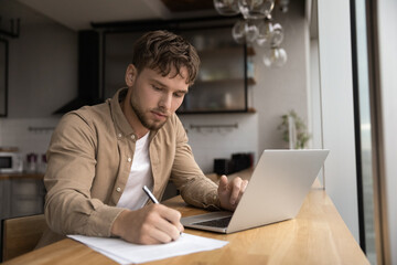 Young guy student prepare research essay at modern kitchen surf internet use laptop write up...