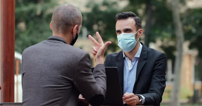 Men in suits and masks on their faces, discussing the prospects of a joint business, sitting at a laptop on the summer terrace of a cafe. The man on the fingers lists the dignity.