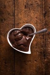 heart shaped bowl filled with chocolate cream with spoon over wooden table. vertical top view