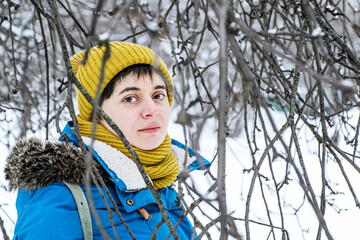 Young woman in bright winter hat and jacket among branches of trees in winter park. Looking into camera. Walks in the winter park, calm mood, sadness. Foreground out of focus
