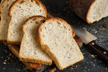 Loaf of fresh sliced white bread is spread out on a cutting board, next to a sharp knife. Close-up of homemade sourdough bread, bread crumbs on black table top