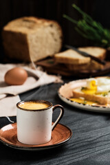 Close-up of cup of espresso coffee with rising steam on black wooden table. On blurred background, soft-boiled egg (poached) in slice of bread, with butter cream and herbs. Breakfast idea