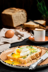 Soft-boiled (poached) egg on slice of bread covered with butter cream and herbs, on clay plate on black wooden table. Espresso coffee and loaf of sliced bread on blurred background. Breakfast idea
