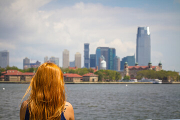 Fototapeta na wymiar A young woman with long red hair is watching the Manhattan skyline from Liberty Island in New York City