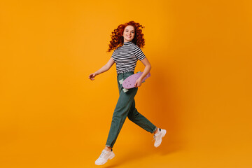 Fototapeta na wymiar Active blue-eyed redhead girl in white stylish sneakers and jeans jumping on orange background, holding lilac longboard