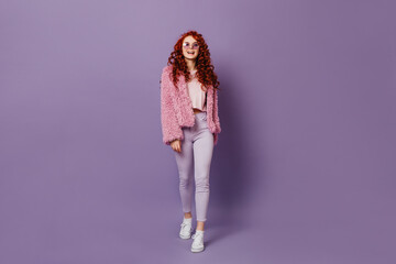 Full-length shot of naughty red-haired girl in round glasses, white jeans and pink coat on purple...