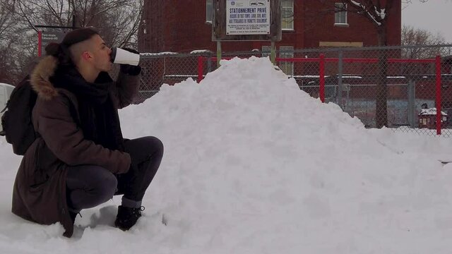 Young man kneeling in snowy park in Rosemont borough of Montreal takes drink of warm coffee or hot chocolate. Guy drinking hot beverage outside in winter.