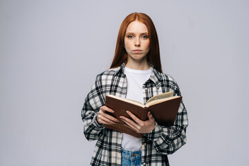 Serious young woman college student holding opened books and looking at camera on isolated gray background. Pretty redhead lady model wearing casual clothes emotionally showing facial expressions