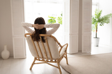 Back view of calm young woman sit rest in cozy modern chair in living room stretch breathe fresh...