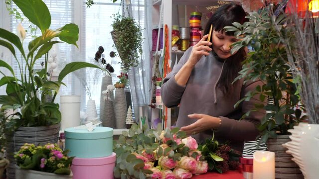 Smiling woman talking by phone in flower shop. Cheerful female florist answering phone call via smartphone and looking at beautiful fresh flowers