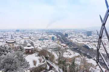 Obraz premium The famous clock tower on Schlossberg hill and historic buildings rooftops with snow, in Graz, Styria region, Austria, in winter day