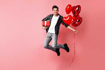 Emotional young man jumping on pink background. Brunette guy with red balloons expressing happiness.