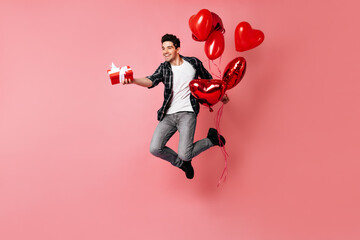 Funny man in jeans jumping on pink background. Dark-haired guy posing with red party balloons and presents.