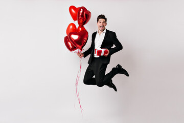 Handsome man in tuxedo jumping with balloons. Indoor photo of smiling guy with gift isolated on white.