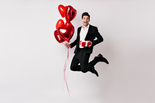 Handsome Man In Tuxedo Jumping With Balloons. Indoor Photo Of Smiling Guy With Gift Isolated On White.