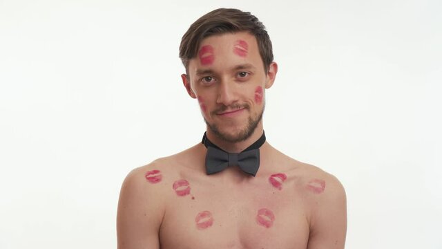 Handsome young brunette man with lipstick kiss marks on body with grey bow tie smile, look at camera isolated on white wall background. Sexy male flirt. Romantic concept of Valentines day close up.