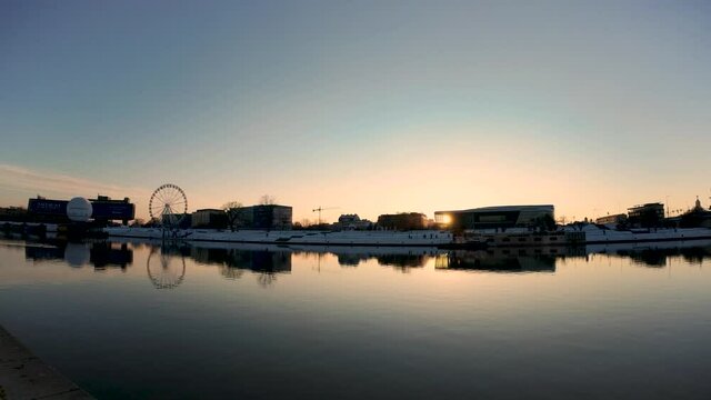 Krakow, Poland - January 31, 2021: Pan shot of vistula river against merry go round and buildings situated in the center of city during sunset. Calm river against sunrise