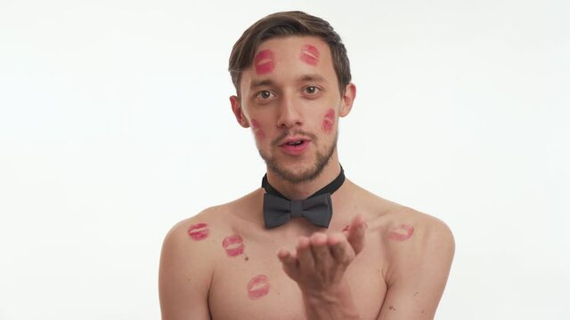 Close up portrait of Caucasian beautiful young brunette bearded man shirtless wear grey bow tie with red lipstick on body and head blowing kiss isolated on white wall background. Women day concept.